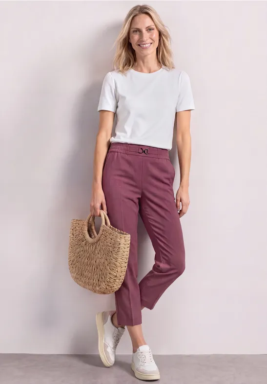 Femme en t-shirt blanc, pantalon violet, baskets blanches et sac en paille devant un fond clair.