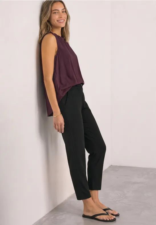 Vrouw leunt tegen een witte muur, draagt een bordeaux top, zwarte broek en sandalen, glimlacht lichtjes.