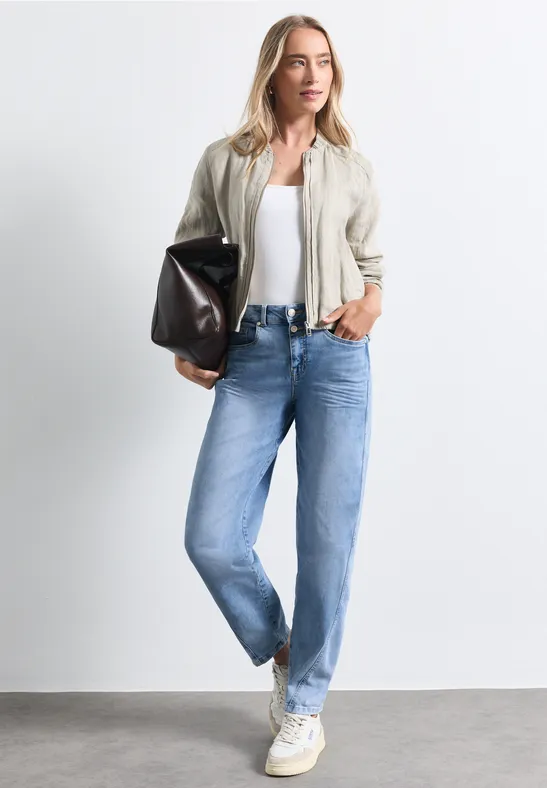 Femme en jean bleu, baskets blanches, top blanc et veste beige, tenant un grand sac noir.