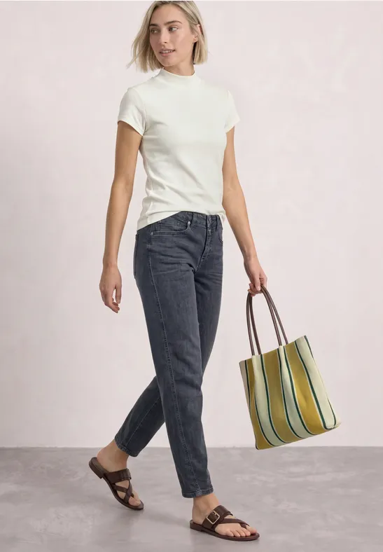 Femme en t-shirt blanc, jean foncé, sandales et sac rayé, posant devant un fond clair.
