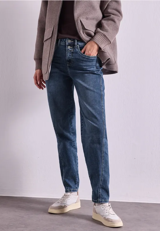 Persoon in casual outfit: grijze overshirt, donkere trui, blauwe jeans en witte sneakers tegen neutrale achtergrond.