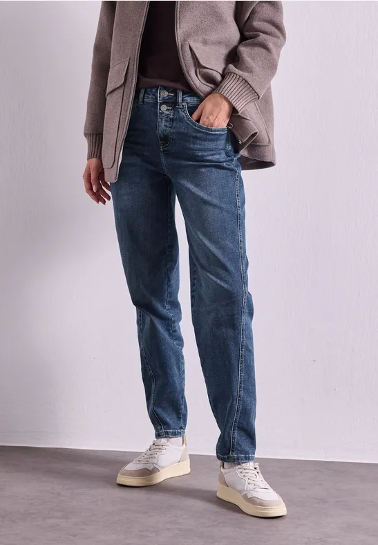 Persoon in blauwe jeans, witte sneakers en jas staat voor een witte muur.