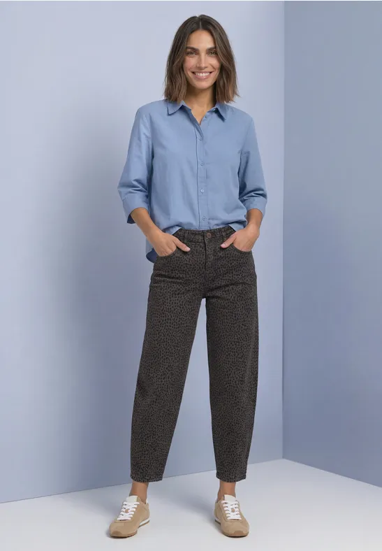 Une femme souriante en chemise bleu clair, pantalon marron à motif et baskets devant un fond bleu pâle.
