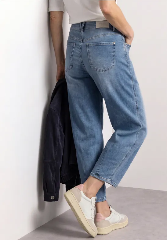 Personne appuyée contre un mur, jean droit, baskets blanches et veste sombre à la main.