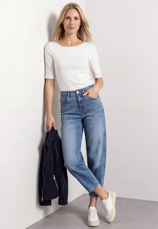 Femme en t-shirt blanc à manches mi-longues, jean et baskets blanches, tenant une veste bleu marine.