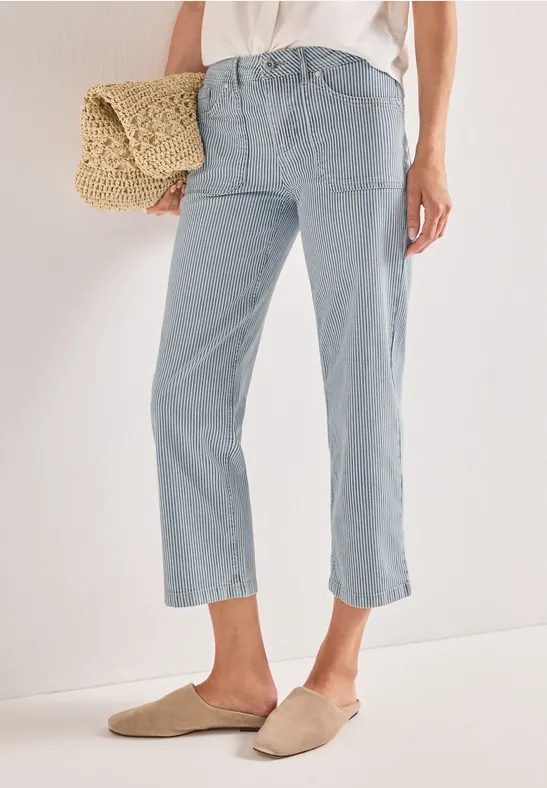 Eine Person trägt hellblaue, knöchelhohe Jeans, beigefarbene Slipper und hält eine Strohtasche.