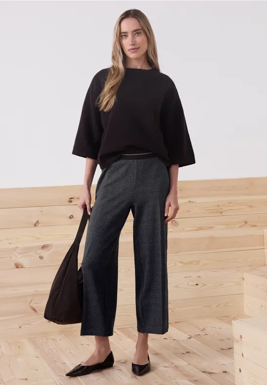 Vrouw in zwarte oversized trui, wijde grijze broek en zwarte ballerina’s, met tas voor lichte houten trap.