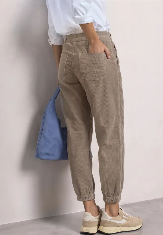 Eine Person trägt beige Cordhose, weiße Schuhe und hält eine blaue Stofftasche.