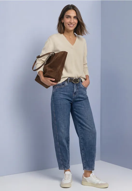 Femme en pull clair et jean, baskets blanches et grand sac devant un fond uni.