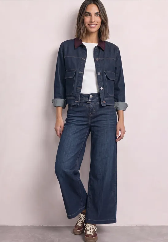 Femme portant une veste en jean, un t-shirt blanc, un jean évasé et des baskets sur fond clair.