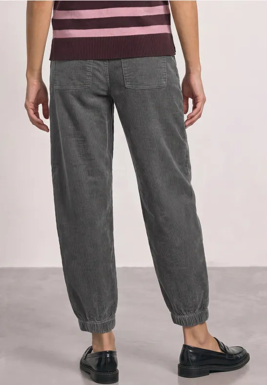 Eine Person von hinten in dunkelgrauen, elastischen Stoffhosen mit Bündchen und schwarzen Slippern.