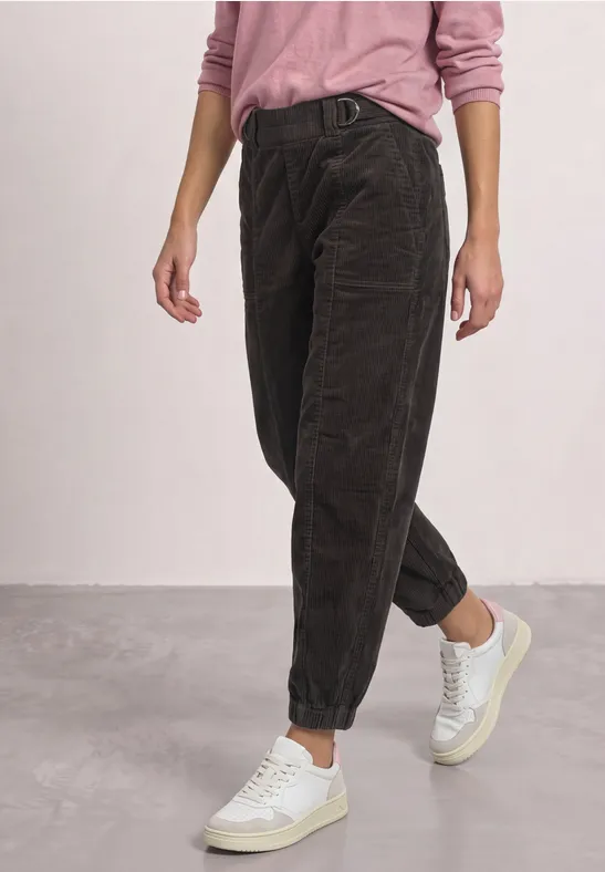 Pantalon en velours côtelé marron foncé, coupe décontractée retroussée, porté avec un pull rose et des baskets blanches.