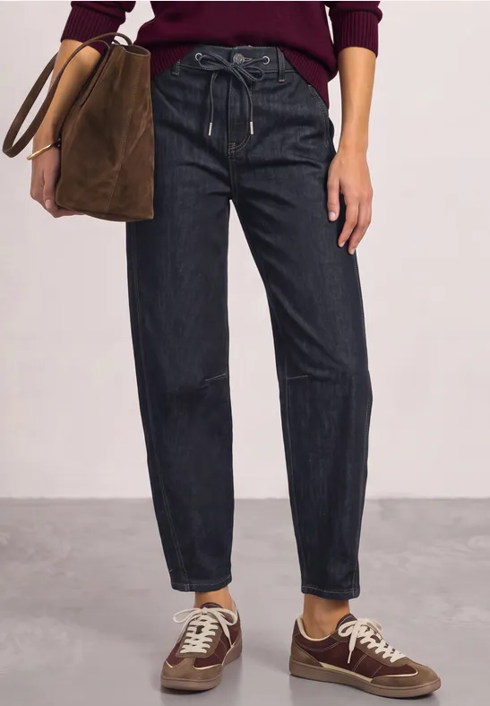 Femme en jean sombre décontracté, baskets brunes, tenant un sac en cuir suédé marron.
