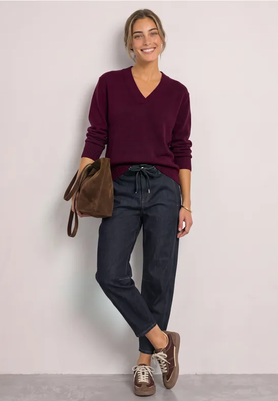 Femme en pull violet à col V, jean foncé et sac marron posant devant un fond neutre.