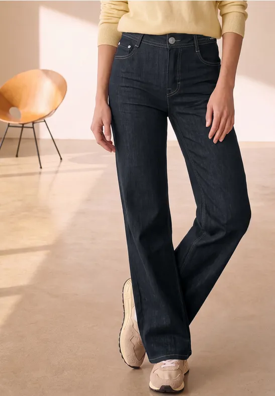 Person trägt dunkle, gerade geschnittene Jeans mit weitem Bein und beigen Sneakers.