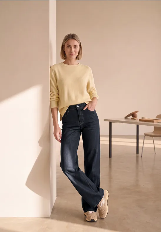 Eine Frau in gelbem Pullover und weiten schwarzen Jeans lehnt sich in einem minimalistischen Raum an die Wand.