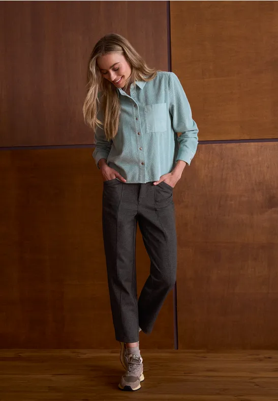 Vrouw leunt tegen houten wand, draagt grijze broek, lichtblouse en hoge hakken, handen in zakken, kijkt omlaag.