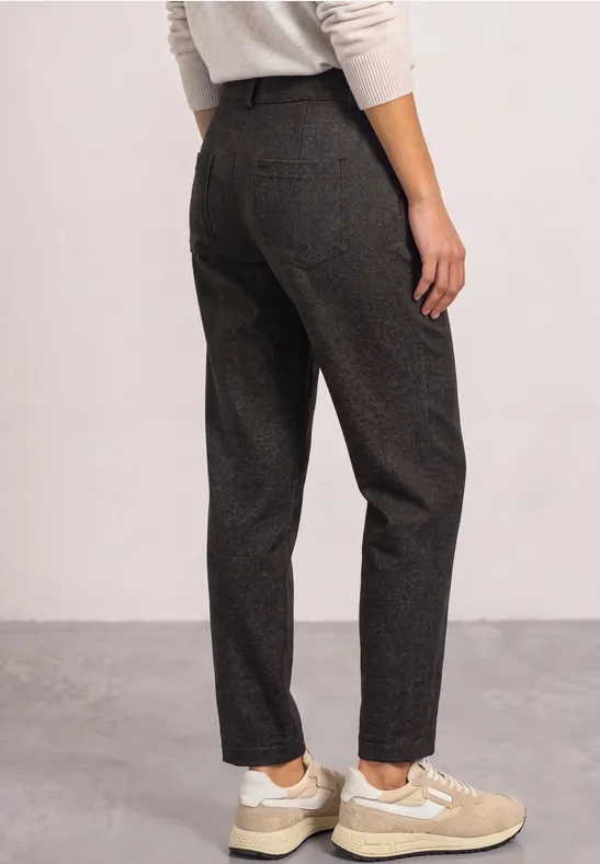 Vrouw van achteren in donkergrijze stoffen broek en beige sneakers tegen neutrale achtergrond.