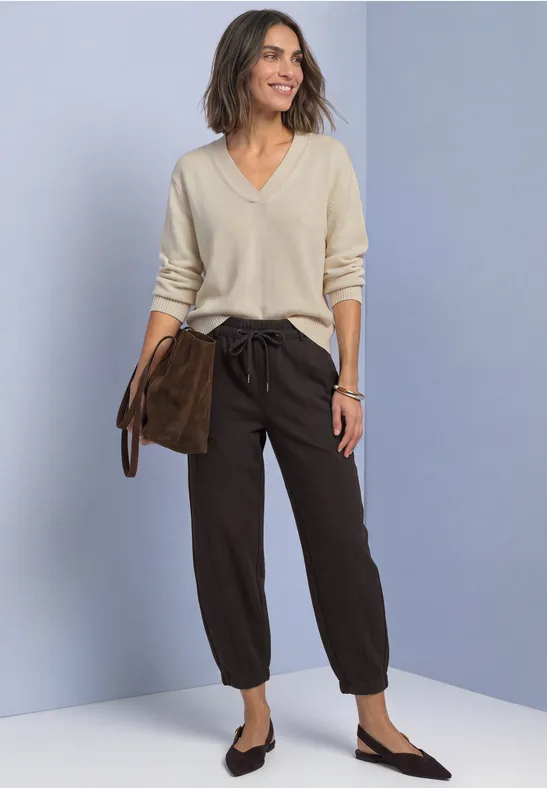 Frau im hellen V-Pullover, lockeren braunen Hosen und schwarzen Loafers, hält eine braune Clutch.