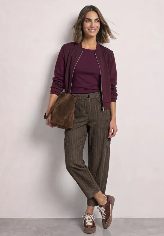 Vrouw in stijlvolle herfstoutfit: paarse top en jas, geruite broek, bruine schoenen en bijpassende tas.
