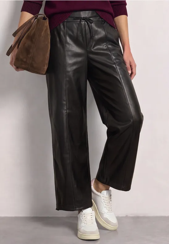 Modischer Look: schwarze, weite Lederhose, weiße Sneaker und braune Wildledertasche vor neutralem Hintergrund.