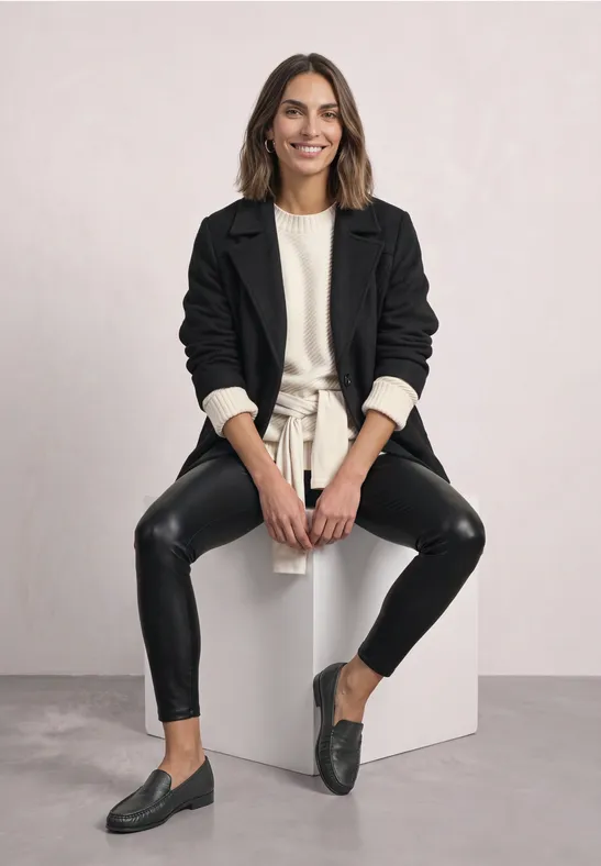 Frau sitzt auf weißem Würfel, trägt schwarzen Blazer, beigen Pullover, schwarze Lederleggings und Loafers.