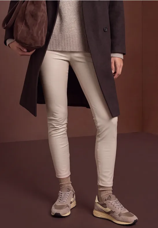 Beine in beigen Lederleggins, grauer Pullover, langer Mantel, Sneaker und große Tasche.