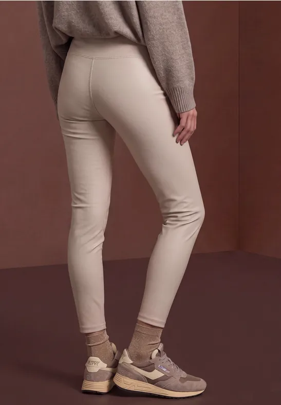 Frau in enger beige Hose und braunen Sneakern vor braunem Hintergrund.