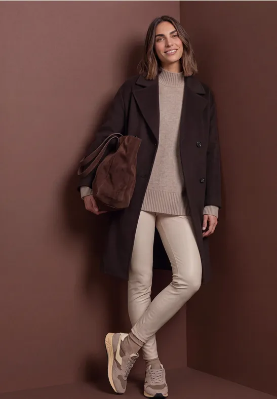Modische Frau in dunkelbraunem Mantel, hellem Pullover, beigen Hosen und Sneakers vor braunem Hintergrund.