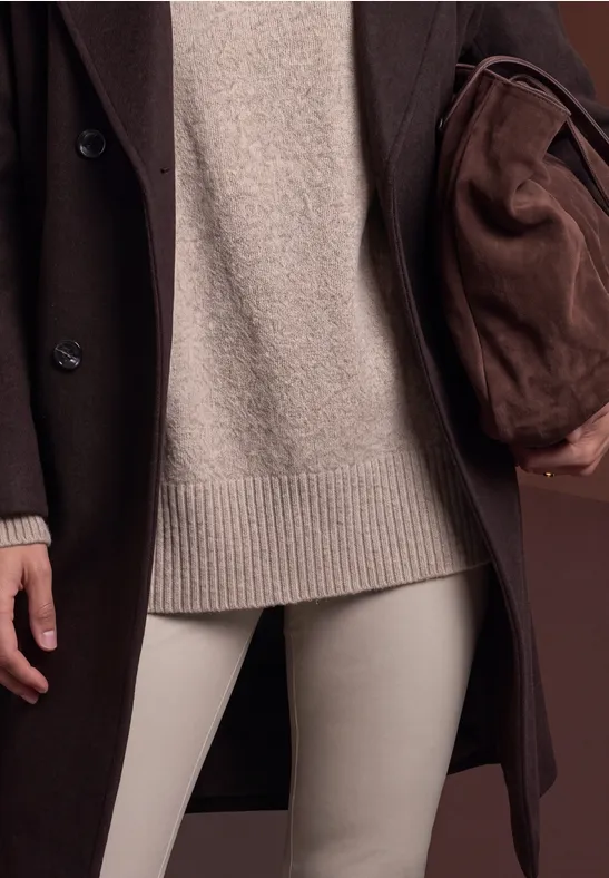 Dunkelbrauner Mantel, beiger Strickpullover, helle Hose und braune Tasche.