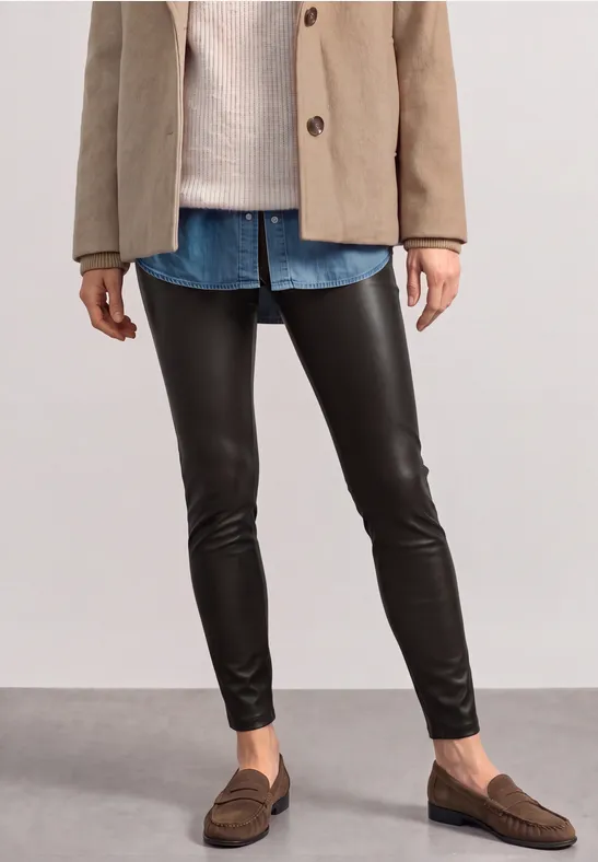 Person trägt schwarze Lederleggings, Jeanshemd, beige Jacke und braune Loafers vor neutralem Hintergrund.