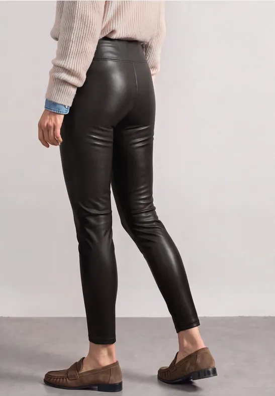 Seitliche Ansicht in schwarzen Lederleggings, beigem Pullover und braunen Loafers vor schlichtem Hintergrund.