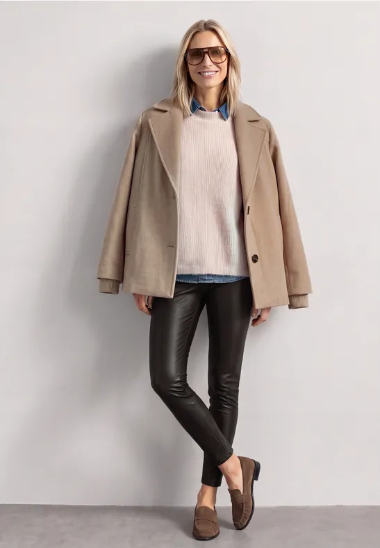 Eine Frau trägt Brille, beigen Mantel, Pulli, Lederleggings und Loafers, lehnt lächelnd an weißer Wand.