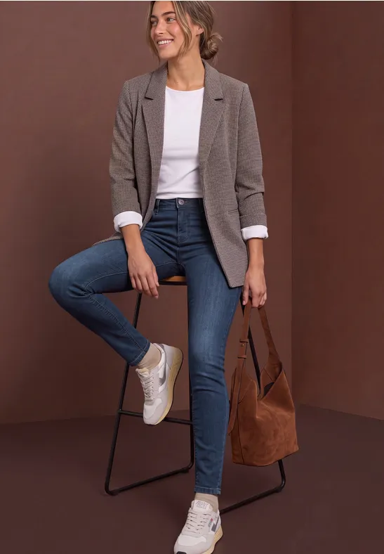 Femme en blazer gris, t-shirt blanc, jean bleu et baskets, assise sur un tabouret avec un grand sac marron.