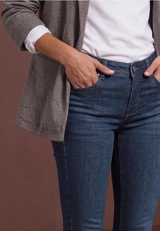 Une personne porte un t-shirt blanc, un blazer à carreaux et un jean bleu, mains dans les poches.