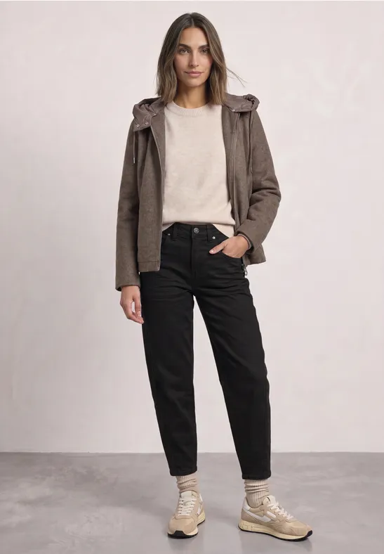 Femme en tenue décontractée: veste marron, pull clair, pantalon noir et baskets beiges sur fond neutre.