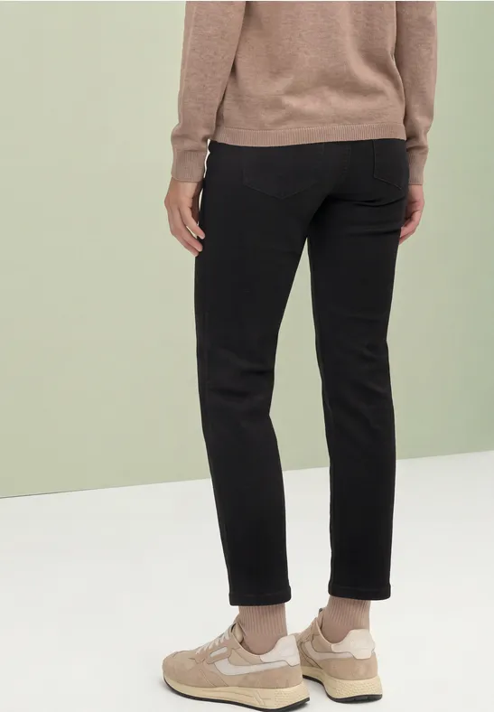 Personne de dos portant un pantalon noir, des baskets beiges et un pull marron sur fond vert et blanc.
