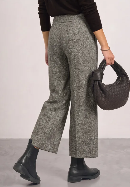 Femme en pantalon gris taille haute, pull noir et bottines, tenant un sac tressé noir.
