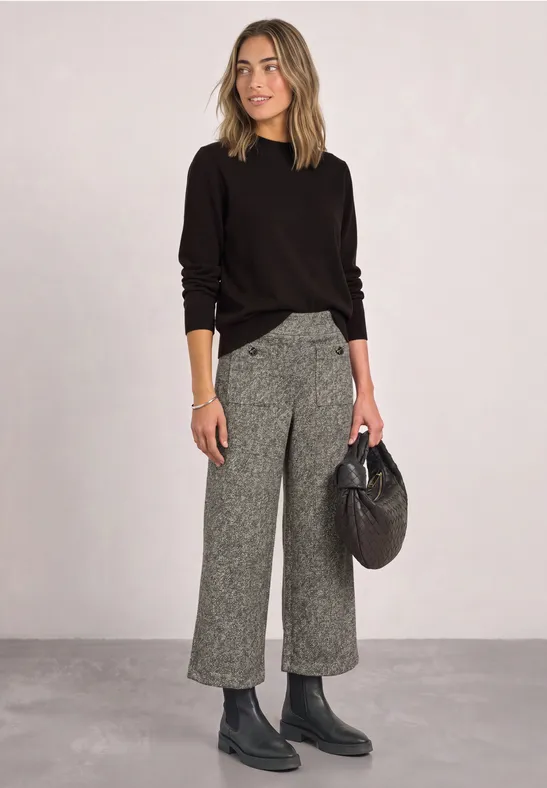 Femme en pull noir, pantalon culotte gris, bottines et sac noir devant un fond neutre.