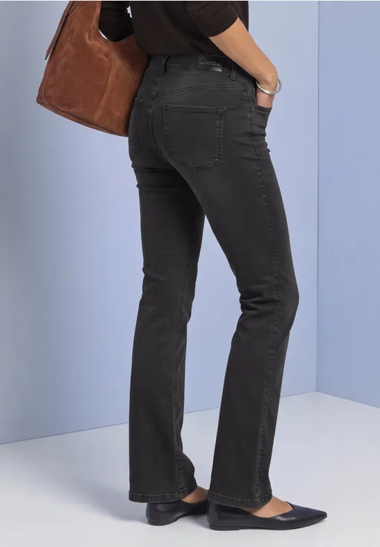 Femme vue de profil portant un jean noir, des ballerines bleu foncé et un sac marron devant un mur bleu clair.