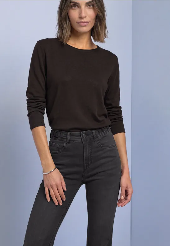 Femme portant un t-shirt à manches longues marron et un jean foncé devant un fond neutre.