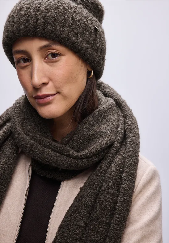 Frau mit brauner Wollmütze und Schal, trägt helle Jacke, neutraler Hintergrund.