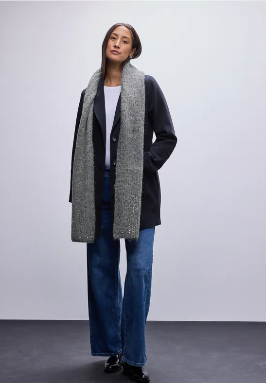 Frau mit grauem Schal, dunklem Mantel und Jeans steht vor hellem Hintergrund.