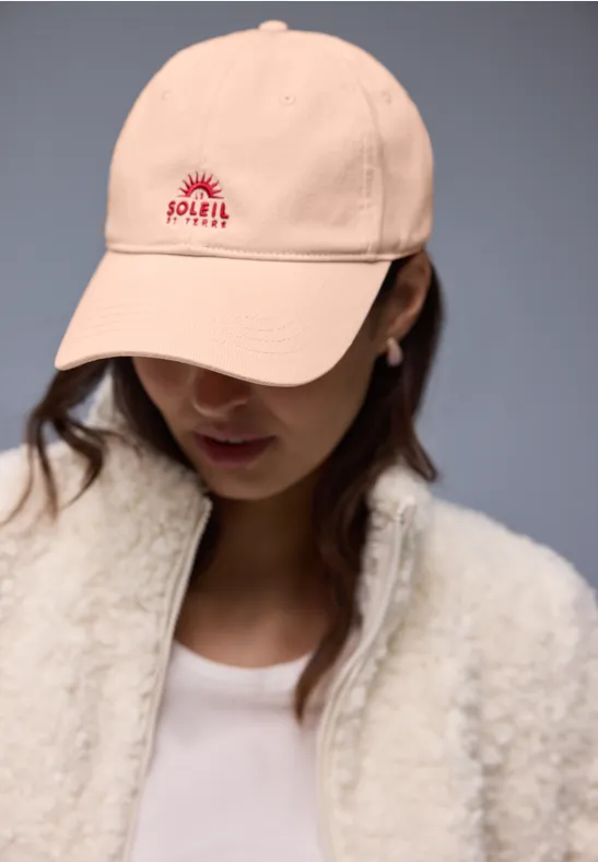 Person trägt beige Cap mit rotem POLLY-Logo, weißem Shirt und flauschiger, cremefarbener Jacke.