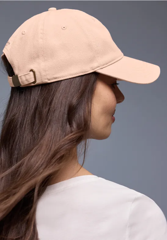 Rückansicht einer Person mit langen braunen Haaren, weißem T-Shirt und beiger Baseballmütze vor grauem Hintergrund.