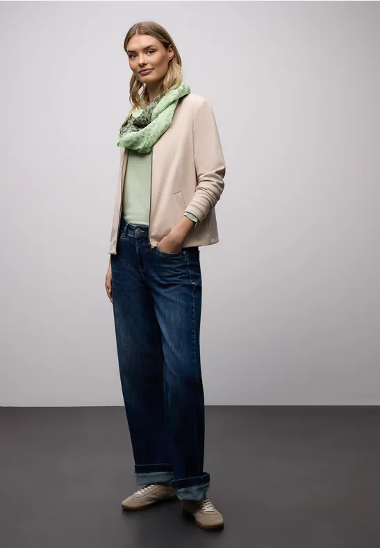 Personne en veste beige, top vert, écharpe assortie, jean large retroussé et mocassins beiges sur fond neutre.