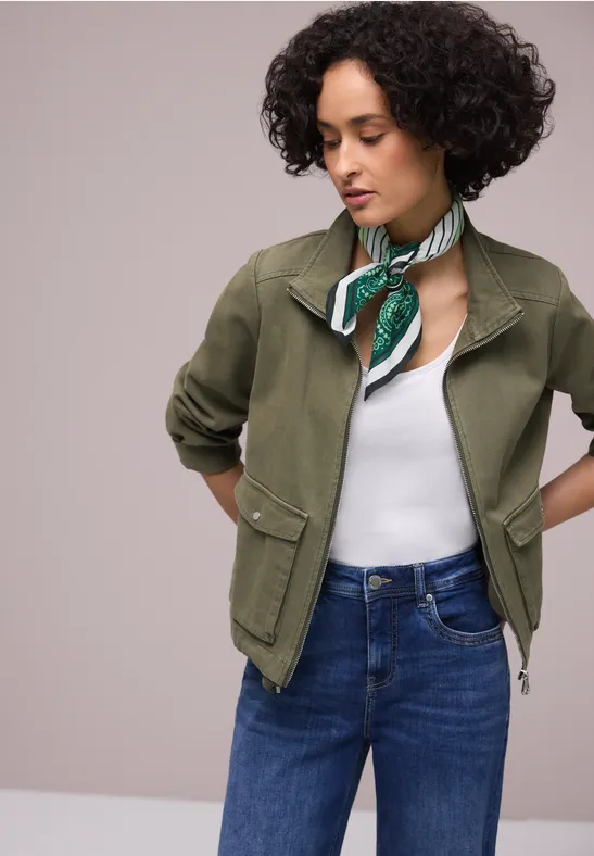 Personne porte une veste olive, un top blanc, un jean bleu et un foulard vert à motifs.