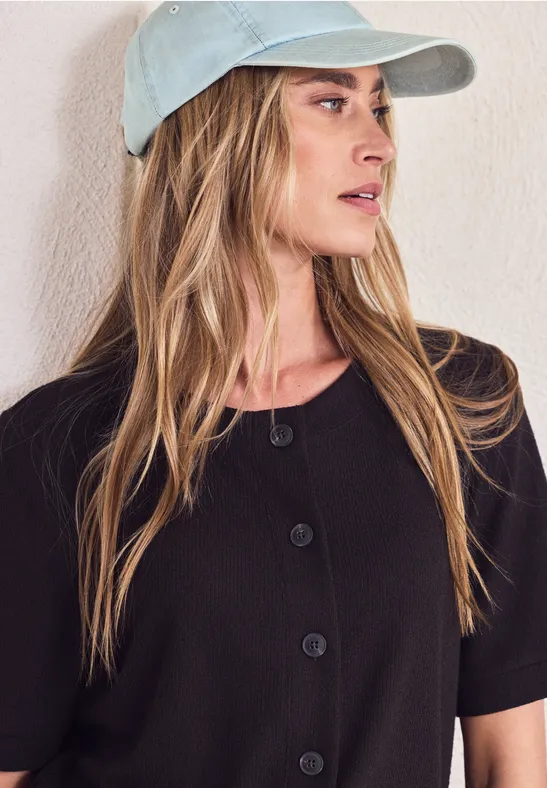 Persoon met pet, lang haar en zwart, knoopbaar shirt, gezicht onscherp.