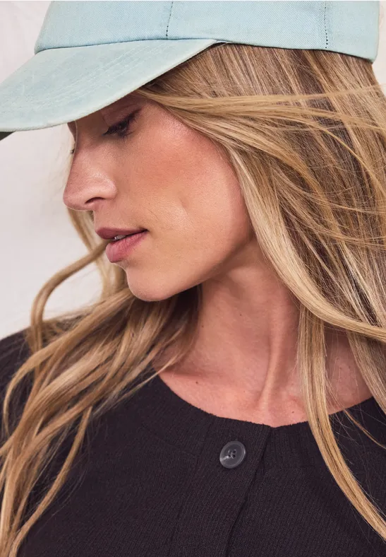 Persoon met lang blond haar, draagt lichtblauwe pet en donkere bovenkleding, gezicht onscherp.