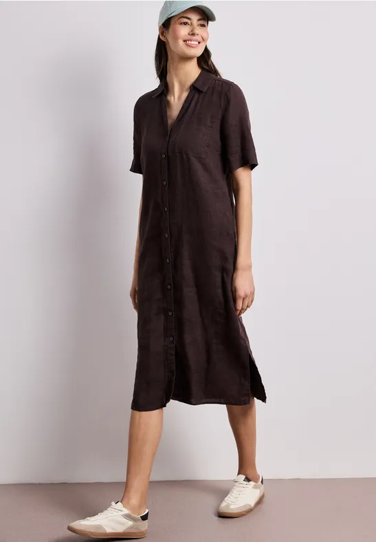 Vrouw draagt een losvallende donkerbruine midi-hemdjurk met korte mouwen, zijsplitsen, beige sneakers en pet.